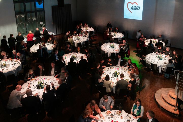 AWO Neujahrsempfang 2023; Impressionen vom AWO Neujahrsempfang, mehrere runde Tiische, die festlich eingedeckt und beleuchtet sind, mit Menschen im Gespräch und am Buffet, das AWO Logo im Hintergrund