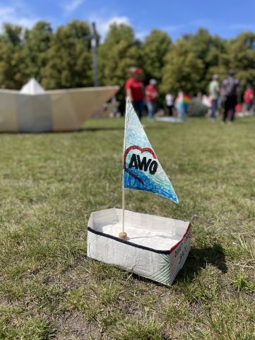 Ein kleines aus Papier gefaltetes Schiffchen mit AWO-Flagge steht auf der Wiese im Hintergrund ein großes Origamiboot und Menschen