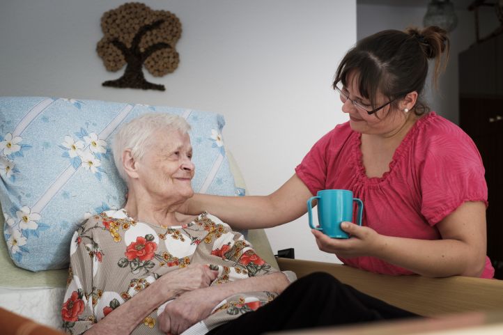 Eine alte Dame Seniorin liegt im Bett und lächelt eine Pflegerin an, die ihr die Hand auf die Schulter gelegt hat und einen Becher in der Hand hält