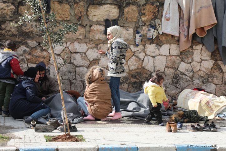 Frauen und Kinder vor einer Häuserwand aus Feldsteinen, Decken hängen an der Wand, Besitztümer und Schuhe liegen auf dem Boden; Foto aus der Erdbebenregion Syrien/Türkei 2023