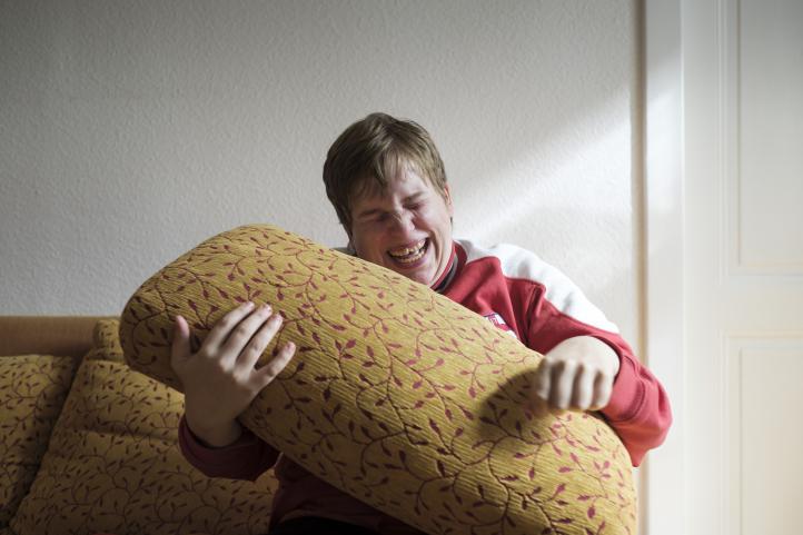 Eine lachende Frau sitzt auf dem Sofa und hält ein Sofakissen über ihren Kopf