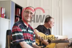 Portraitaufnahme lächelnder Mann mit Brille und Halbglatze hinter ihm ein Mann mit Gitarre 