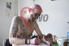 Ein älterer Mann mit Tattoos sitzt mit einem Brötchen am Tisch und löst ein Kreuzworträtsel