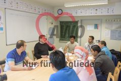 Sechs Männer und eine Frau mit Kopftuch sitzen an einem Tisch in einem Klassenzimmer und hören dem Lehrer zu, der etwas erklärt