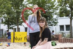 Zwei junge Frauen sind auf einem Spielplatz mit Kindern und lächeln in die Kamera