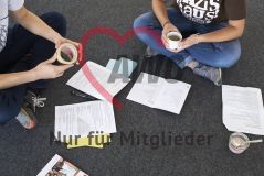 Zwei junge Menschen mit Klebeband und einer Teetasse sitzen auf dem Boden in einem Seminar Workshop und auf dem Boden liegen Papiere Arbeitsmaterialien