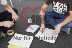 Zwei junge Menschen mit einem Stift sitzen auf dem Boden in einem Seminar Workshop und auf dem Boden liegen Papiere Arbeitsmaterialien