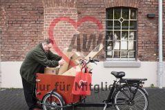  Mann an einem mit Paketen beladenem Lastenrad, am Lenker ein roter AWO-Beutel