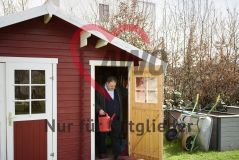   Mann mit roter Gießkanne an der Tür eines roten Gartenschuppens, daneben Schubkarren