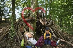 zwei Kinder im Wald, vor einem Tipi aus Ästen beim Picknick sitzend
