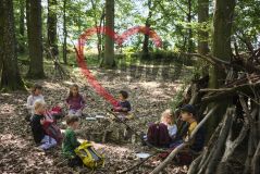 sieben Kinder im Wald, vor einem Tipi aus Ästen beim Picknick sitzend