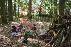 sechs Kinder im Wald, vor einem Tipi aus Ästen beim Picknick sitzend