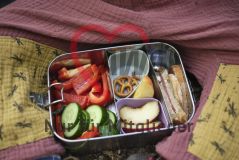 Brotdose (mit Gemüse, Obst, Brot und Minibrezeln) zwischen Kinderbeinen